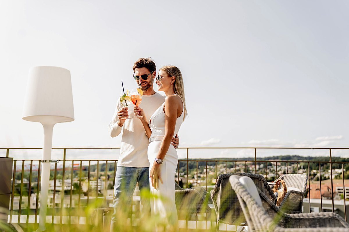 Ein Paar steht auf der Rooftop Bar Terrasse mit einem Cocktail in der Hand geniesst die Aussicht