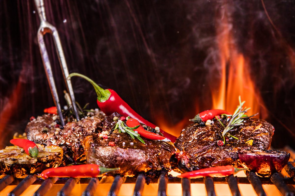 Grillgabel trifft Fleisch und Chili auf einem Grillrost und im Hintergrund flackert das Feuer
