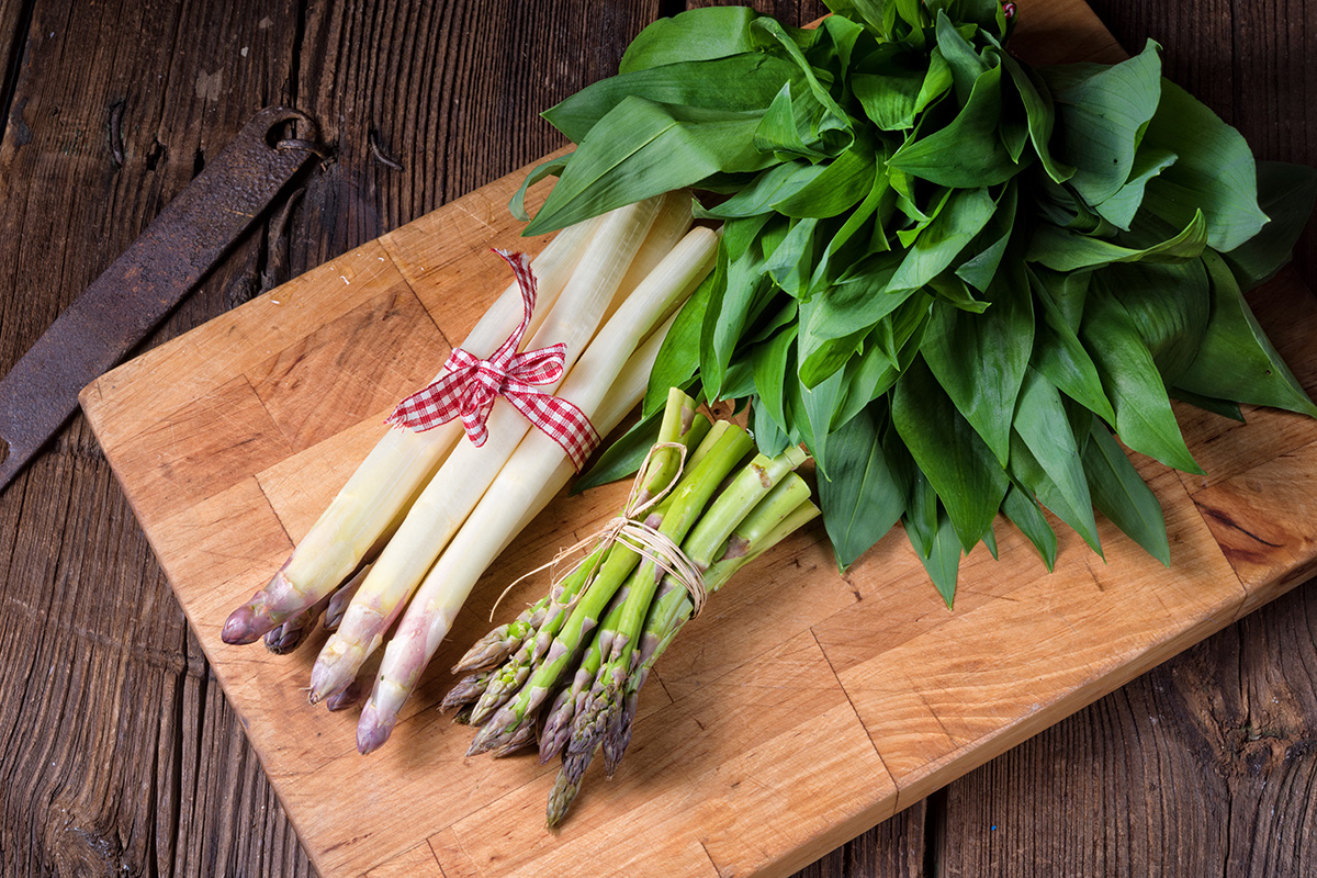 Frühlingshafte Nahaufnahme von weissem und grünem Spargel sowie frischem Bärlauch auf einem rustikalen Holzbrett.