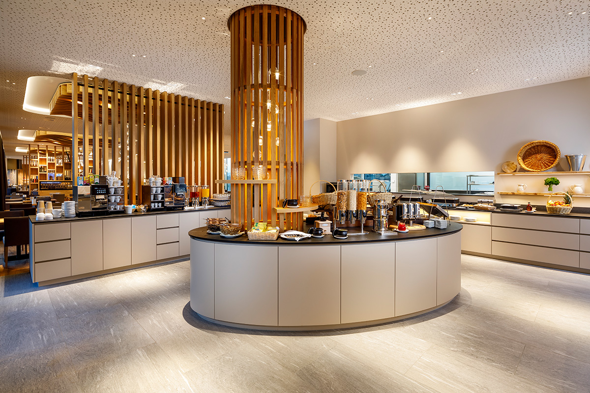 Modernes Frühstücksbuffet in der Allegra Lodge Kloten mit zentraler Holz-Säule, Kaffeestation, Müsli-Auswahl und warmen Speisen in hellem, alpinem Design.