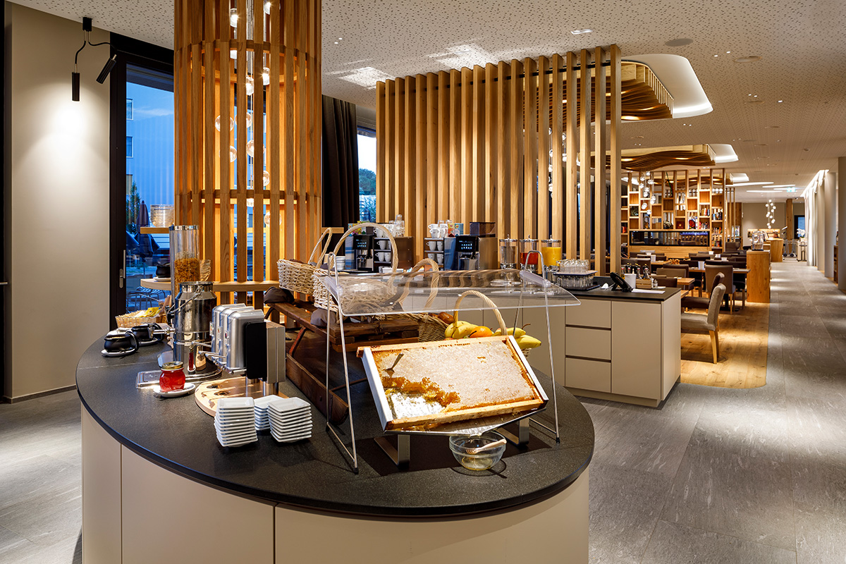 Frühstücksbuffet im Restaurant Tschadun mit frischer Honigwabe im Vordergrund, Brotauswahl und Blick in den modernen, gemütlichen Gastraum mit viel Holz.