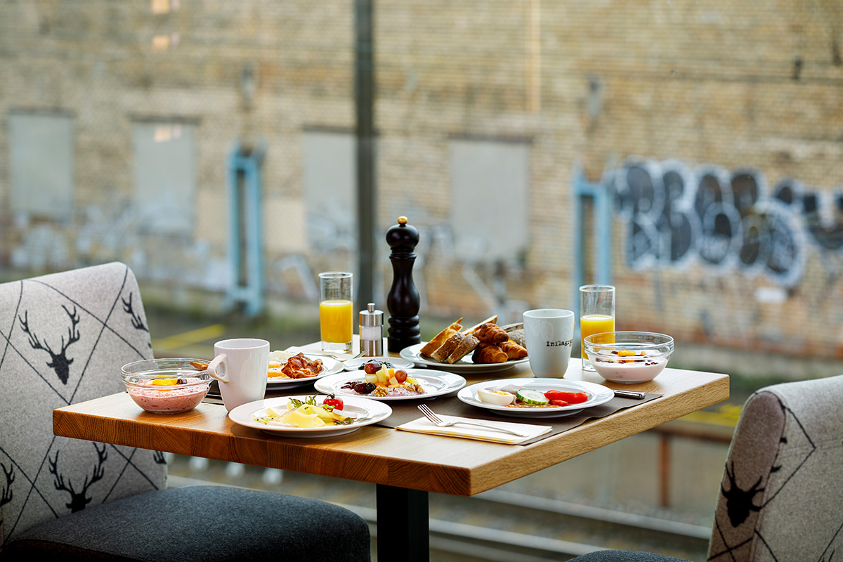 Reichhaltig gedeckter Frühstückstisch im Restaurant Tschadun mit Blick durch ein Fenster auf eine urbane Backsteinfassade. Zu sehen sind frische Eierspeisen, Säfte, Gebäck und Sessel mit Hirsch-Motiv im Lodge-Stil.